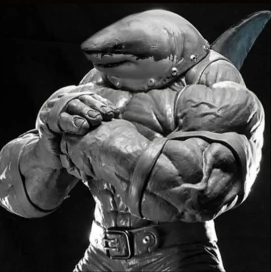 The image shows a muscular humanoid shark. It has the head of a shark, with a large mouth and sharp teeth. Its body is muscular and defined, with large pectoral muscles, arms, and legs. The shark is wearing a pair of pants and has a belt around its waist. Its skin is gray and its eyes are black. The shark is standing in a powerful pose, with its fists clenched and its arms crossed in front of its chest. It looks like it is ready to