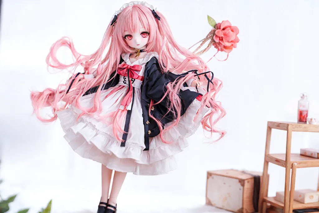 The image shows a doll with pink hair and red eyes. She is wearing a white and pink dress with a black jacket. She is also wearing a black bow tie and a pink rose in her hair. She is standing on a white table and there is a pink rose bush next to her. The background is white.