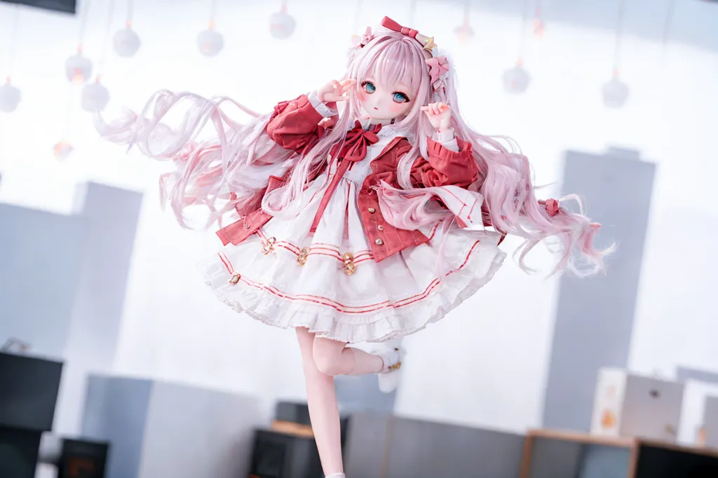 This is an image of a doll with long pink hair, blue eyes, and a red and white dress. The doll is standing on one leg and has her arms in the air. She is wearing a pink bow in her hair and has a big smile on her face. The background is white and there are some small, round lights in the background.