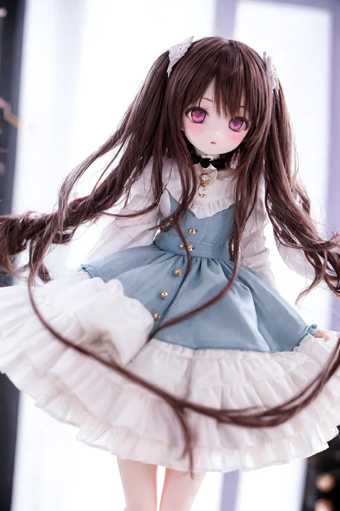 The image shows a doll with long brown hair, purple eyes, and a light blue dress with white ruffles. The doll is standing on a white background and is looking at the camera. The doll's hair is styled in two pigtails with white bows. The doll's dress has gold buttons and a white collar. The doll's shoes are white with pink bows. The doll's skin is pale and flawless. The doll's eyes are bright and shiny. The doll's lips are slightly parted. The doll's expression is serene and peaceful.