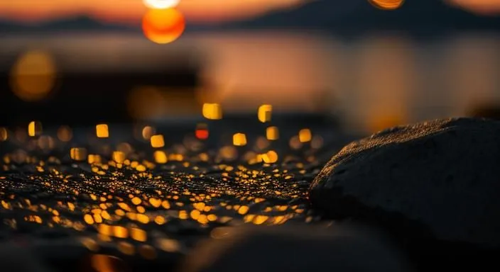 The setting sun casts a golden glow on the wet rocks and pebbles along the shore. The warm colors and soft focus create a peaceful and tranquil scene. The sun is setting behind a mountain, casting a warm glow over the water. The sky is a deep blue, and the water is a dark blue. The only light comes from the sun and the stars. The scene is peaceful and serene.